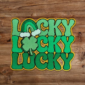Puede incluir: Una alfombra de bienvenida verde y amarilla con la palabra "LUCKY" repetida tres veces en una fuente retro. Un trébol y un arcoíris se muestran encima de la palabra superior.