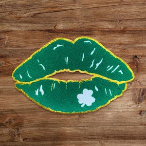 May include: Green glitter lips with a white shamrock in the center, on a brown wooden background.
