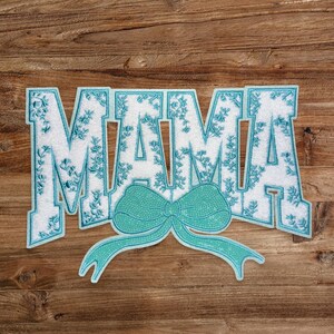 May include: A white felt patch with the word "MAMA" in blue outlined letters with a blue and white floral pattern. A blue bow is attached to the bottom of the word.