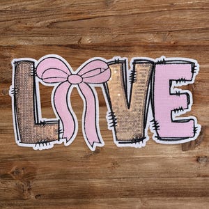 May include: A pink and gold felt applique of the word "LOVE" with a pink bow in the center of the "O".