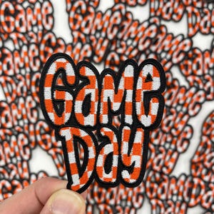 May include: A close-up of a red and white checkered fabric patch with the words "Game Day" embroidered in black. The patch is surrounded by other patches with the same design.