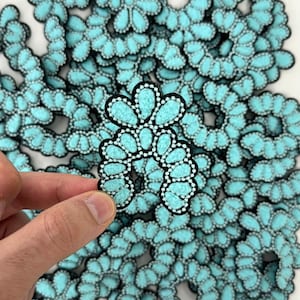 May include: A collection of turquoise felt appliques with a floral design. Each applique features a black border, white dotted accents, and a unique petal-like shape. The appliques are ideal for crafting and embellishment projects.