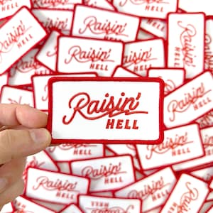 May include: White rectangular patch with a red border and the embroidered text "Raisin' HELL" in red cursive. The patch is held in a hand, with many similar patches scattered in the background.