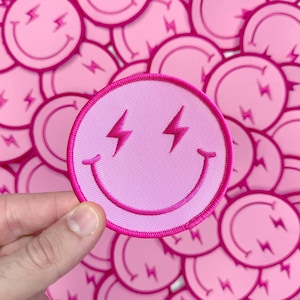 May include: Pink smiley face patch with lightning bolt eyes. The patch is surrounded by other pink smiley face patches with lightning bolt eyes.