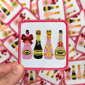May include: Embroidered patch featuring four champagne bottles in pink, black, and beige with gold accents. The patch has a white background and a pink border. The design includes red bows and decorative labels.
