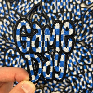May include: A close-up of a blue and white checkered fabric patch with the text "Game Day" in a bold, black font.