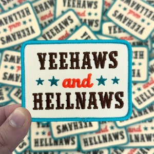 May include: A rectangular patch with the words "YEEHAWS and HELLNAWS" in brown, with the word "and" in red, and blue stars. The patch has a light blue border and is held in front of a pile of similar patches.