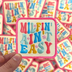 May include: A square embroidered patch with the text "MILFIN' AIN'T EASY" in colorful, bubble-style letters. The patch has a cream-colored background, a pink border, and is surrounded by similar patches.