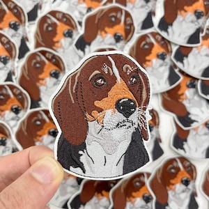 May include: A close-up of a brown, white, and black embroidered patch of a beagle dog's face. The patch is surrounded by other similar patches.