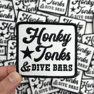 May include: A white patch with black embroidered text that reads "Honky Tonks & Dive Bars" with a black star in the center. The patch is surrounded by other similar patches.