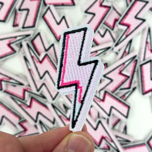 May include: A pink and black embroidered lightning bolt patch. The patch has a white border and a pink center with a black outline. A bright pink line runs down the center of the lightning bolt. Other patches are in the background.