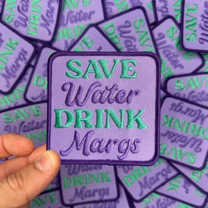 May include: A close-up of a stack of purple and green embroidered iron-on patches that say "Save Water Drink Margs".