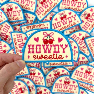 May include: A collection of iron-on patches with a light yellow background, blue trim, and pink embroidery. The patches feature the text "HOWDY sweetie" with two cherries and hearts.