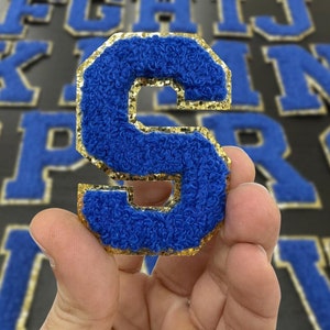 May include: A blue felt letter S with gold glitter trim. The letter is held in a hand.
