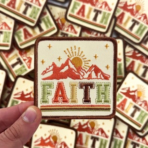 May include: A beige fabric patch with a brown border. The patch features an embroidered image of a mountain range with a sun and stars. The word "FAITH" is embroidered in colorful letters below the mountain range.