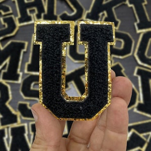 May include: Black felt letter U with gold glitter trim. The letter is held in a hand against a background of other letters.
