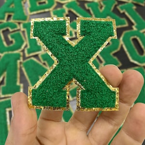 May include: Green felt letter X with gold glitter trim. The letter is held in a hand.
