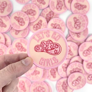 May include: A collection of pink and peach-colored embroidered patches. One patch features a cowboy hat with star details and the text "LET'S GO GIRLS" in pink thread. The patches are round and have a stitched border.