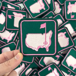 May include: A collection of green and pink embroidered patches. Each patch features a pink outline of the United States with a golf flag and pin, set against a white background. The patches have a pink border.
