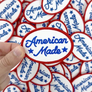 May include: Oval-shaped embroidered patches with the text "American Made" in blue script and stars, surrounded by a red border. The patches are white with a blue script font. A hand holds one patch, with many more in the background.