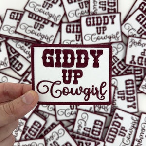 May include: A close-up of a white patch with the text "Giddy Up Cowgirl" in maroon embroidery. The patch is surrounded by other similar patches with different text.
