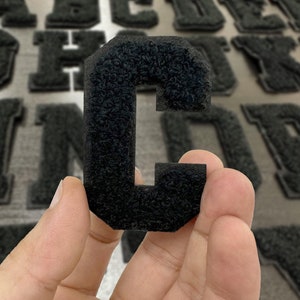 May include: A black felt letter C, the letter is thick and fuzzy, the letter is being held by a hand.