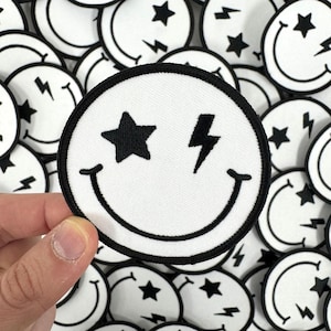 May include: A white embroidered patch with a black smiley face, a star, and a lightning bolt. The patch is surrounded by other similar patches.