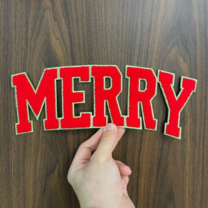 May include: A red felt applique spelling "MERRY" with a green border, held against a wood grain background. The letters are bold and blocky, perfect for holiday crafts and decorations.
