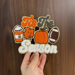 May include: A decorative patch with the words "Tis The Season" and fall-themed images. The patch features a coffee cup, leaf, pumpkin, and football, all in orange, brown, and white with gold glitter accents.