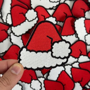 May include: Red and white Santa hat patch with a black border. The patch features a textured red hat with a white fluffy trim. The patch is held up by a hand, with many more patches in the background.