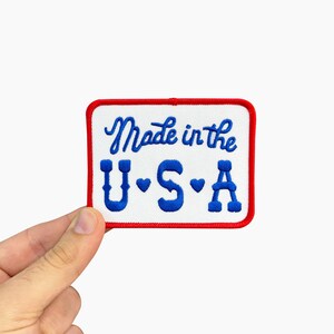 May include: Embroidered patch with a white background and a red border. The patch features the text "Made in the U.S.A." in blue script and block letters. The patch is rectangular with rounded corners.