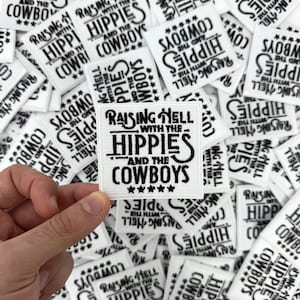 May include: White embroidered patch with the text "Raising Hell with the Hippies and the Cowboys" in black. The patch has a square shape with a black border and five stars at the bottom. Other similar patches are in the background.