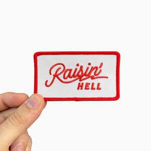 May include: Embroidered patch with the words "Raisin' Hell" in red script on a white rectangular background. The patch has a red border and is held by a hand against a white background.