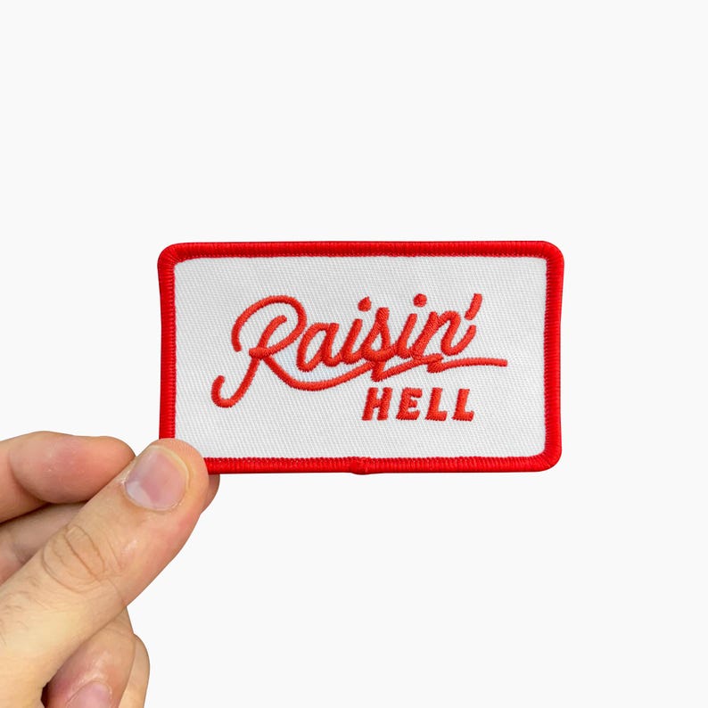 May include: Embroidered patch with the words "Raisin' Hell" in red script on a white rectangular background. The patch has a red border and is held by a hand against a white background.