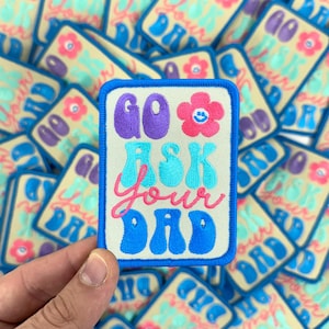 May include: A stack of colorful iron-on patches with a blue border and a white background. The patch in the front reads "Go Ask Your Dad" in a retro font with a pink flower with a smiley face in the center.