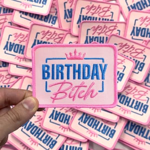 May include: A stack of pink iron-on patches with the text "Birthday Bitch" in blue and pink lettering. The patches have a pink border and a small crown design.