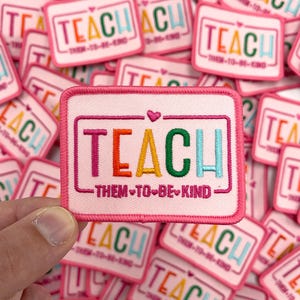 May include: Pink fabric patch with the word "TEACH" in rainbow colors and the text "THEM TO BE KIND" in black with a heart between each word.