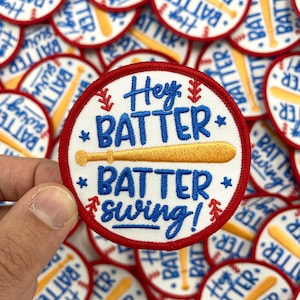 May include: A close-up of a white, round, embroidered patch with red stitching. The patch features a baseball bat with a yellow handle and a blue and white text that reads "Hey Batter Batter Swing!"