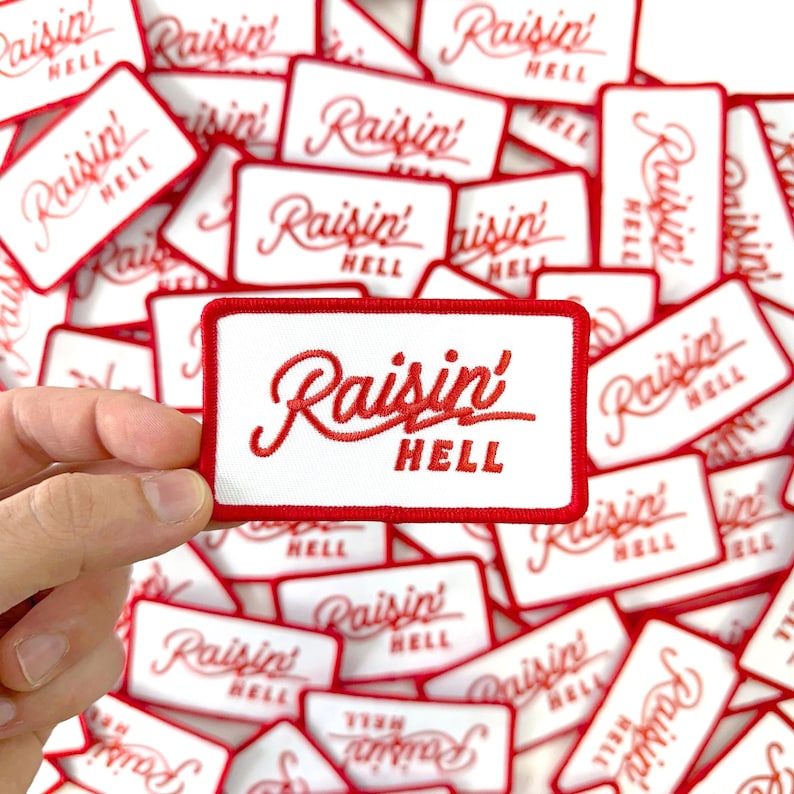 May include: White rectangular patch with a red border and the embroidered text "Raisin' HELL" in red cursive. The patch is held in a hand, with many similar patches scattered in the background.