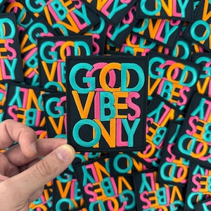 May include: A close-up of a hand holding a black patch with the words "GOOD VIBES ONLY" in colorful, overlapping letters. The patch is surrounded by other similar patches, all with the same phrase but in different color combinations.
