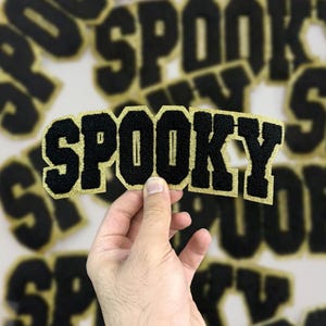 May include: Black and gold "Spooky" letter patches. The patches have a black, textured font with a gold glitter border. The word "Spooky" is displayed in a bold, collegiate-style font. Other patches are visible in the background.