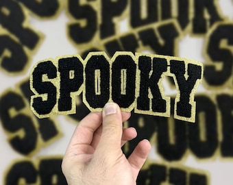 Può includere: Toppe con lettere "Spooky" nere e dorate. Le toppe hanno un carattere nero strutturato con un bordo glitterato dorato. La parola "Spooky" è visualizzata in un carattere in grassetto in stile collegiale. Altre toppe sono visibili sullo sfondo.