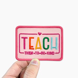 a hand holding a patch that says teach them to be kind