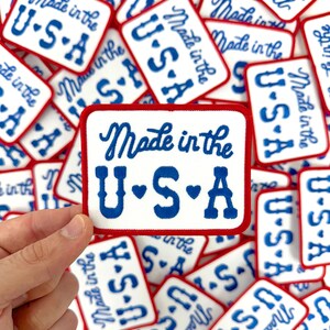 May include: Rectangular white patches with a red border and blue text reading "Made in the U.S.A." The patches are scattered, with one held by a hand. The text is in a cursive font.