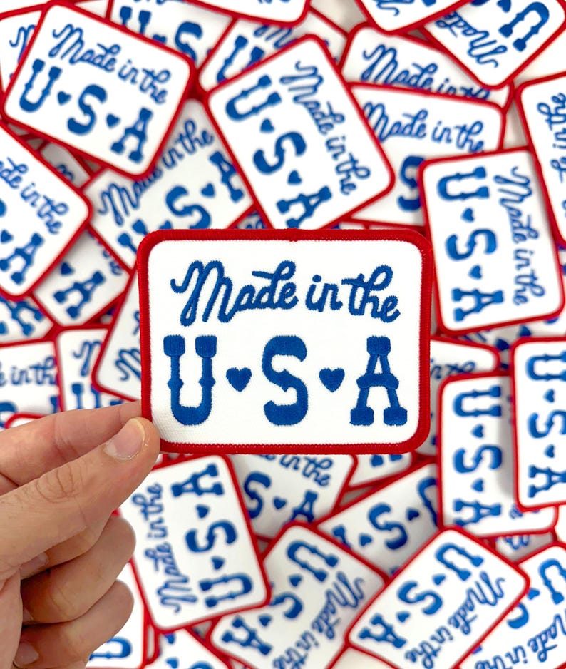 May include: Rectangular white patches with a red border and blue text reading "Made in the U.S.A." The patches are scattered, with one held by a hand. The text is in a cursive font.
