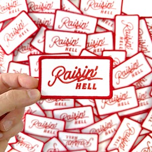 May include: Rectangular white patch with a red border and the embroidered text "Raisin' HELL" in a cursive font. The image shows a hand holding one patch, with many similar patches scattered in the background.