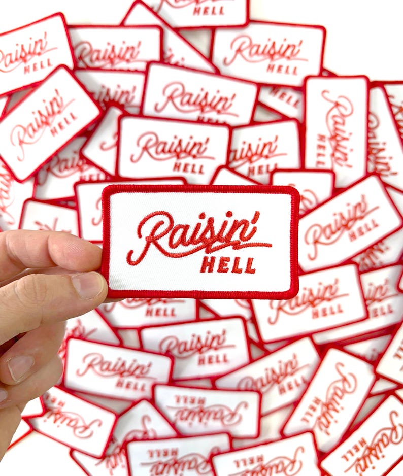 May include: Rectangular white patch with a red border and the embroidered text "Raisin' HELL" in a cursive font. The image shows a hand holding one patch, with many similar patches scattered in the background.