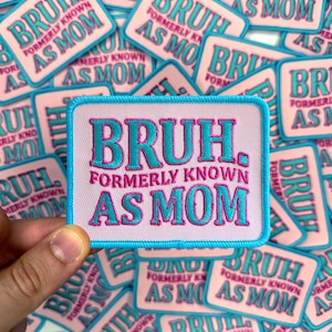 May include: A collection of pink embroidered patches with the text "BRUH, FORMERLY KNOWN AS MOM" in teal and pink. The patches have a light blue border. The patches are rectangular and are scattered across the image.