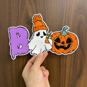 May include: A Halloween-themed patch featuring the word "BOO" in purple, a white ghost wearing an orange beanie and holding a drink, and an orange jack-o'-lantern. The patch is held up by a hand.