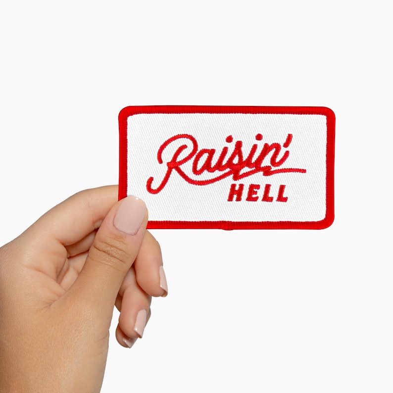 May include: Embroidered patch with a white background and a red border. The patch features the words "Raisin' HELL" in red script and block letters. The patch is rectangular in shape.
